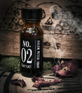 An Apothecary bottle of Vampfangs Fragrance: No.2 Salem Witch Shop photographed in an actual Salem Witch Shop named Hex. A cracked, wooden board with an etched in skull. Smoking rose petals and some green moss complete the photo as accents.