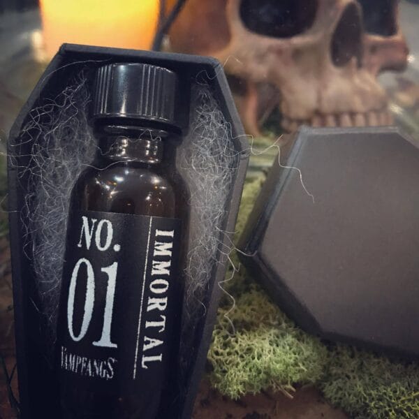 An Apothecary bottle of Vampfangs Fragance: Immoral #1 with a Black Handmade Coffin Box - Background set of human skull and a glowing candle.